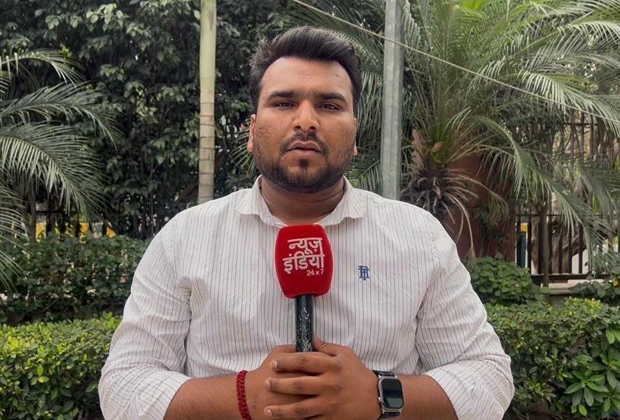 Male reporter in a white shirt holds a red news microphone outdoors, with palm trees in the background.