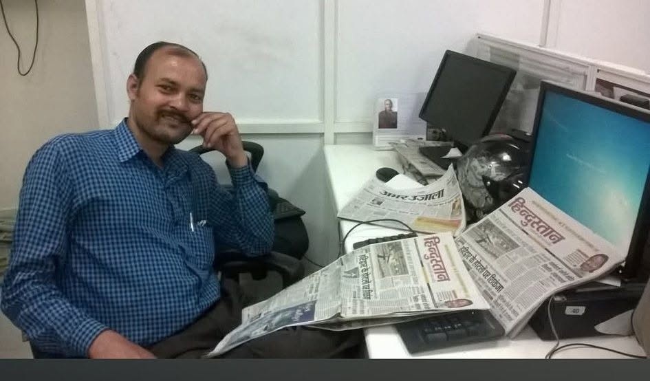 khushal goswami | No. 1 Indian Media News Portal Man in a blue checkered shirt sits at a cluttered office desk with newspapers spread across the desk and two computer monitors in the background, smiling at the camera.