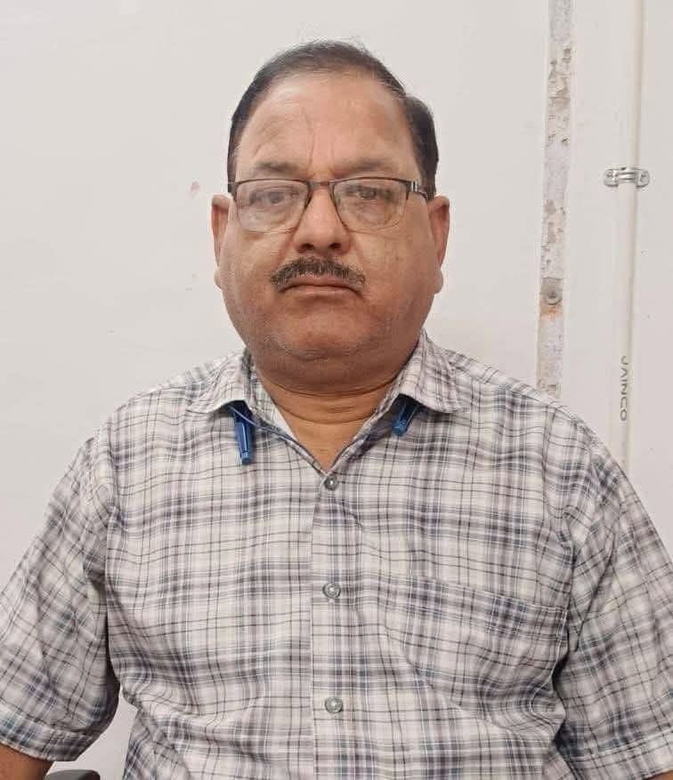 Middle-aged man with glasses wearing a plaid shirt sits facing the camera indoors with a neutral expression.