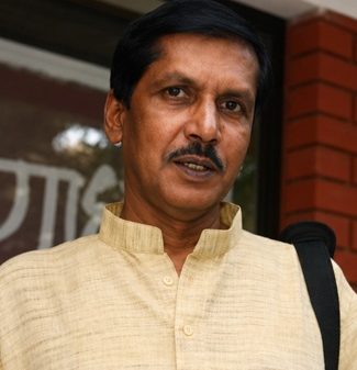Portrait of a man with a mustache wearing a light yellow kurta, standing outdoors with a shoulder bag strap visible.