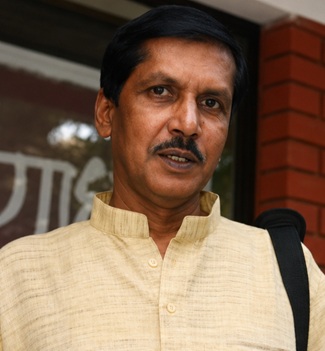 Portrait of a man with a mustache wearing a light yellow kurta, standing outdoors with a shoulder bag strap visible.