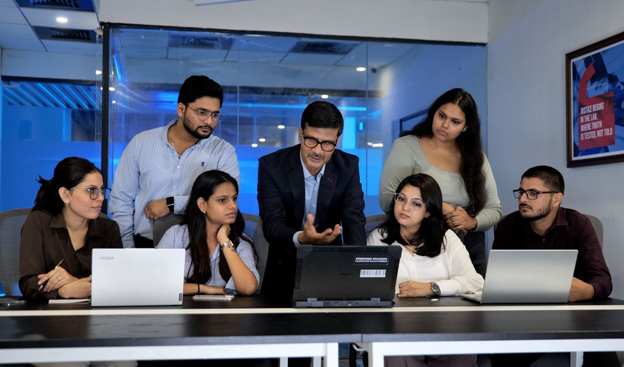 Seven colleagues in a glass-walled office gather around a conference table, with a man in a suit gesturing toward a device while others look on and use laptops.