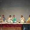 Six musicians seated cross-legged on a stage, playing Indian classical instruments (tabla, flutes) with microphones in front of them, a backdrop screen behind.