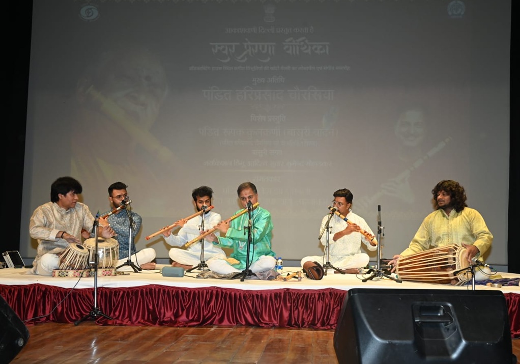 Six musicians seated cross-legged on a stage, playing Indian classical instruments (tabla, flutes) with microphones in front of them, a backdrop screen behind.