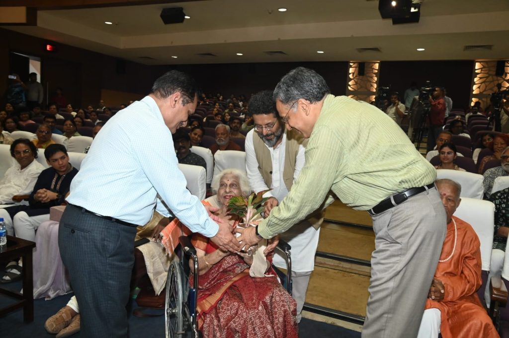 lokarpan_1 | No. 1 Indian Media News Portal Elderly woman in wheelchair receiving a small plant from two men at a formal event in an auditorium.