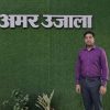 A man in a purple shirt stands against a green textured wall with large white Hindi letters above him.