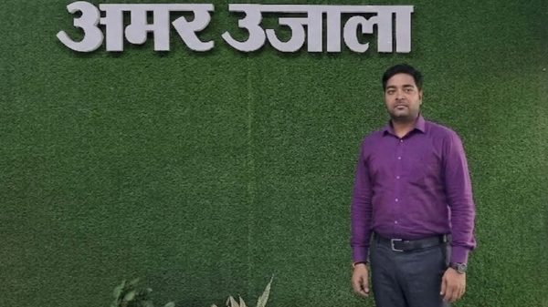 A man in a purple shirt stands against a green textured wall with large white Hindi letters above him.