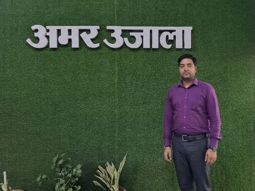 A man in a purple shirt stands against a green textured wall with large white Hindi letters above him.