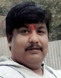 Close-up of a man with a red tilak on his forehead, outdoors near a corrugated metal fence.