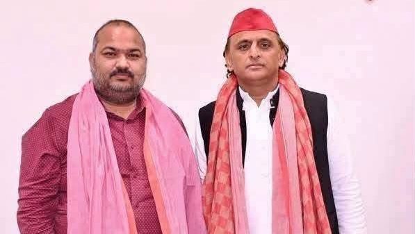 Two men pose for a photo indoors, both wearing pink scarves; one in a maroon shirt, the other in a white kurta with a red head wrap.