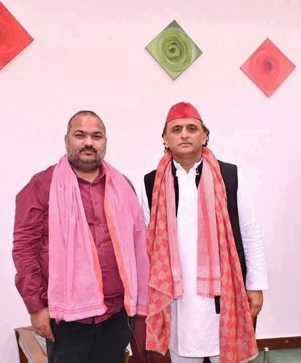 Two men pose for a photo indoors, both wearing pink scarves; one in a maroon shirt, the other in a white kurta with a red head wrap.