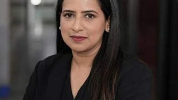 Professional head-and-shoulders portrait of a woman with long dark hair in a black blazer, looking at the camera in an office setting