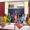 Eleven women in colorful Indian attire pose behind a table with name placards at a community event.