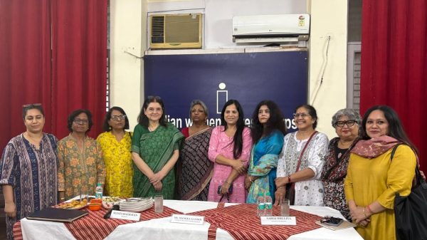 Eleven women in colorful Indian attire pose behind a table with name placards at a community event.