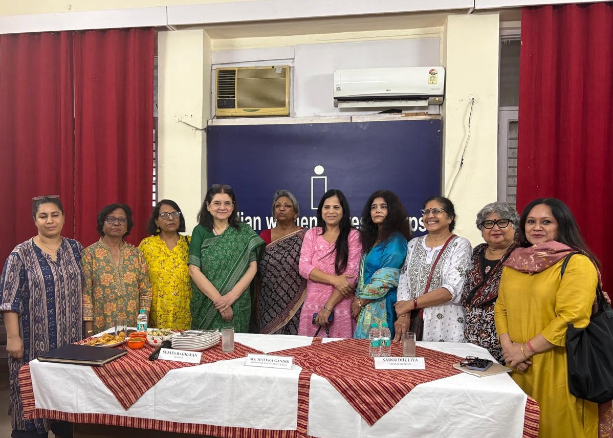 Eleven women in colorful Indian attire pose behind a table with name placards at a community event.