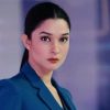 Professional portrait of a woman with dark hair tied back, wearing a blue blazer, looking at the camera with a neutral expression in a blurred indoor setting.