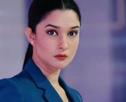 Professional portrait of a woman with dark hair tied back, wearing a blue blazer, looking at the camera with a neutral expression in a blurred indoor setting.