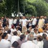 Large group of people gathered outdoors in a park, many seated on the ground with fists raised, suggesting a protest or demonstration amid trees and onlookers standing nearby.