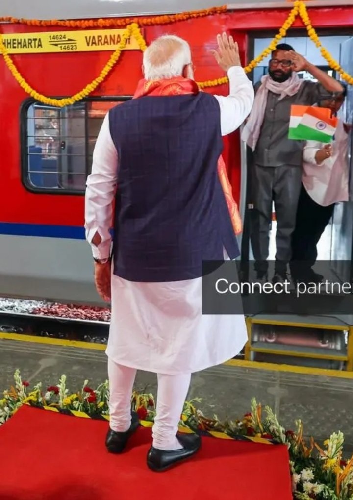 modi train | No. 1 Indian Media News Portal Older man in a white kurta and dark waistcoat waves from a red train car decorated with yellow marigold garlands; a man on the train holds an Indian flag.