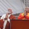 Two elderly Indian men stand at a podium on a stage, hands joined in prayer, looking upward in a devotional moment.
