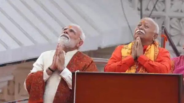 Two elderly Indian men stand at a podium on a stage, hands joined in prayer, looking upward in a devotional moment.