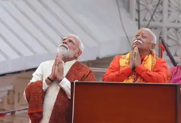 Two elderly Indian men stand at a podium on a stage, hands joined in prayer, looking upward in a devotional moment.