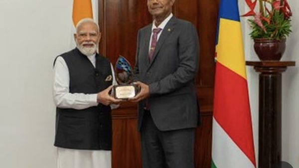 Two men in formal attire exchange a glass award in a ceremonial setting, with national flags in the background.