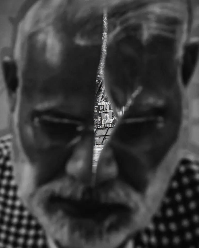 modi_nyt | No. 1 Indian Media News Portal Close-up of an older man's face seen through a narrow vertical crack, with a distant political poster in view.
