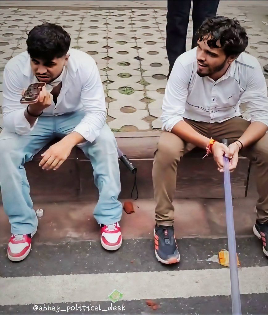 msf | No. 1 Indian Media News Portal Two young men sit on a curb outside; left man in a white shirt and light blue pants holds a phone to his mouth while the right man in a white shirt and tan pants smiles, gripping a long purple tube.