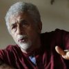 Older man with gray hair and beard, wearing a maroon shirt, gesturing with his hand as he speaks indoors to someone off-camera.
