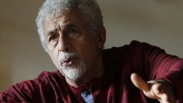 Older man with gray hair and beard, wearing a maroon shirt, gesturing with his hand as he speaks indoors to someone off-camera.