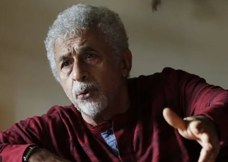 Older man with gray hair and beard, wearing a maroon shirt, gesturing with his hand as he speaks indoors to someone off-camera.