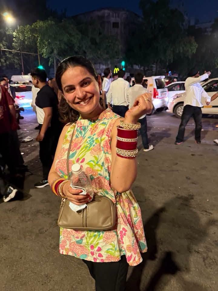 navvivahita | No. 1 Indian Media News Portal Smiling woman in a colorful floral sleeveless top poses on a busy street at night, holding a water bottle and a beige purse, with people and cars in the background.