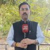 Male news reporter outdoors holding a red microphone with 'न्यूज़ इंडिया 24x7' text, wearing glasses and a dark Nehru jacket over a light shirt, with trees in the background.