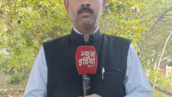 Male news reporter outdoors holding a red microphone with 'न्यूज़ इंडिया 24x7' text, wearing glasses and a dark Nehru jacket over a light shirt, with trees in the background.