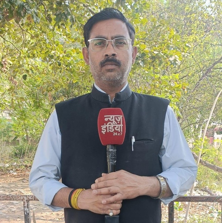 Male news reporter outdoors holding a red microphone with 'न्यूज़ इंडिया 24x7' text, wearing glasses and a dark Nehru jacket over a light shirt, with trees in the background.