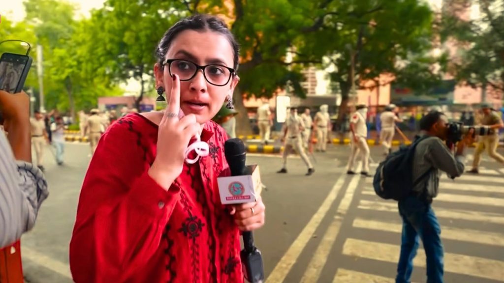 neha | No. 1 Indian Media News Portal Reporter in red with glasses speaks into a handheld mic on a city street, police officers and pedestrians in the background.