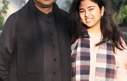 Man in a black outfit stands beside a young woman in a plaid scarf outdoors; bottom news banner reads about Dharmendra Pradhan's daughter, Instagram, and online backlash over US LLM studies.