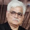 Portrait of an older man with white hair and glasses, wearing a black shirt, looking at the camera.