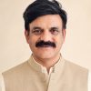 Portrait of a middle-aged man with dark hair and a mustache, wearing a beige Nehru jacket against a light background.