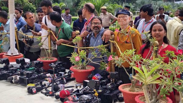 Group of reporters and spectators behind a rope barrier, facing a table of cameras and microphones outdoors among potted plants and greenery.