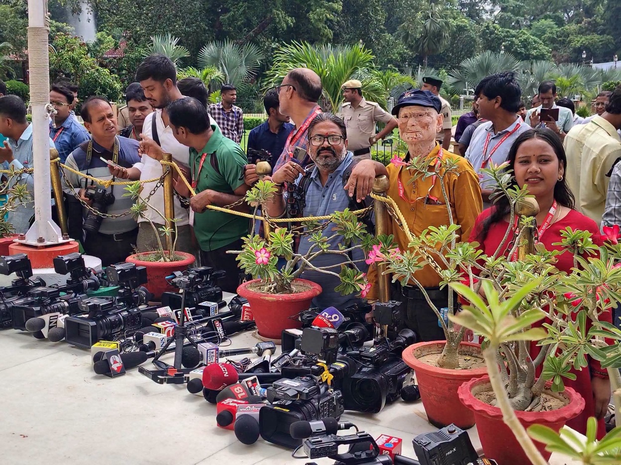 Group of reporters and spectators behind a rope barrier, facing a table of cameras and microphones outdoors among potted plants and greenery.