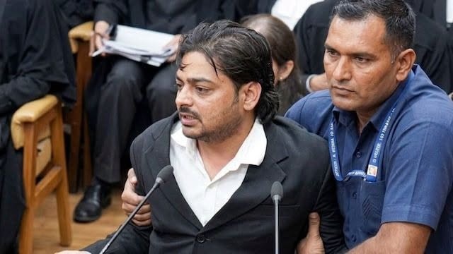 Man in a dark suit speaks into two microphones while another man supports him in a courtroom setting.