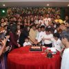 Large group of people gathered around a red table cutting a birthday cake with candles lit, celebrating together.