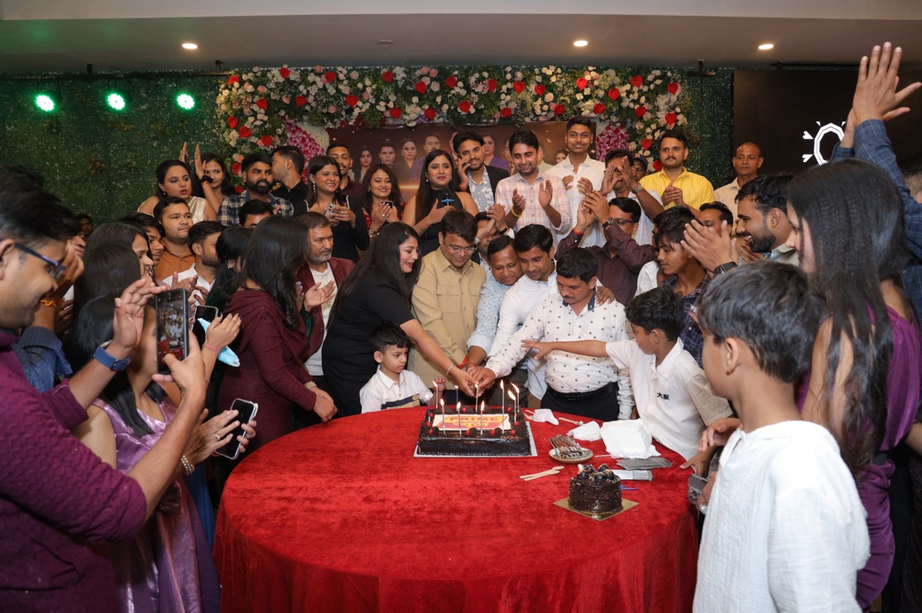 Large group of people gathered around a red table cutting a birthday cake with candles lit, celebrating together.