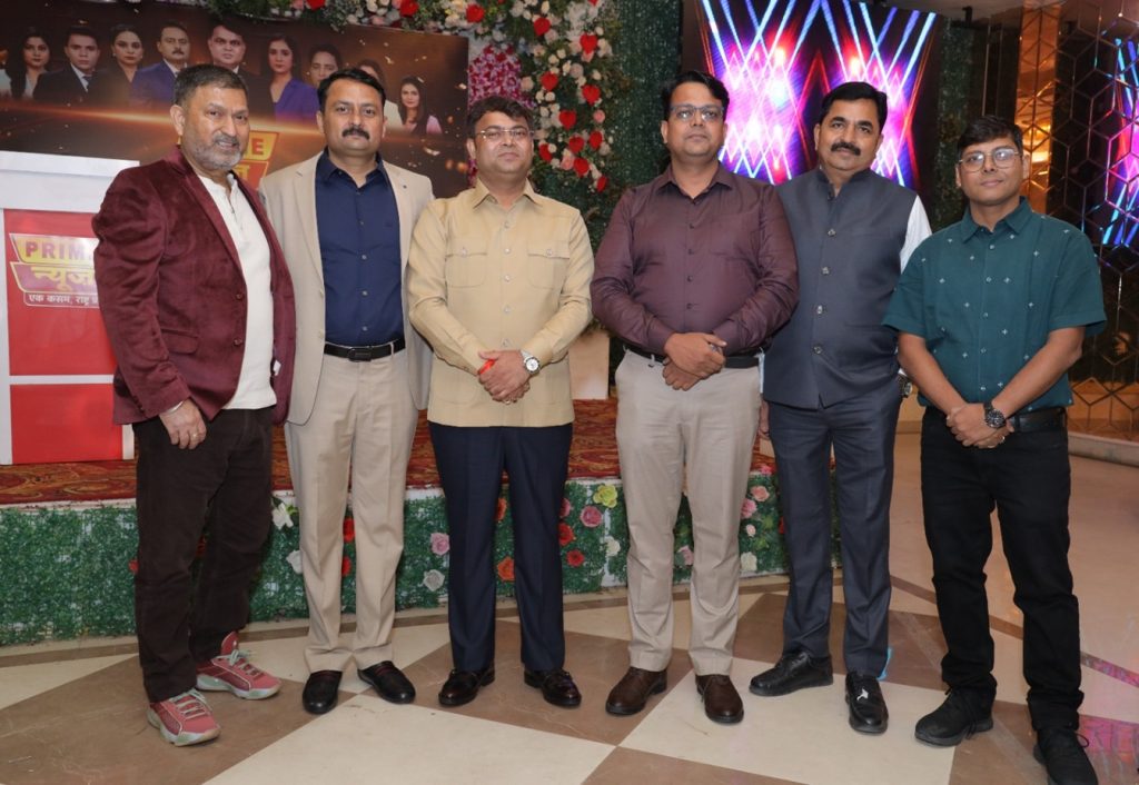 prime news_1 | No. 1 Indian Media News Portal Six men in formal attire posing for a group photo at an indoor event with a floral backdrop and colorful stage lights on a decorated stage.