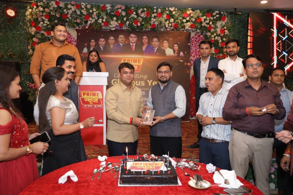 prime news_2 | No. 1 Indian Media News Portal Colleagues celebrate at a decorated event around a chocolate cake, presenting a gift in front of a 'Prime' banner and flower backdrop.