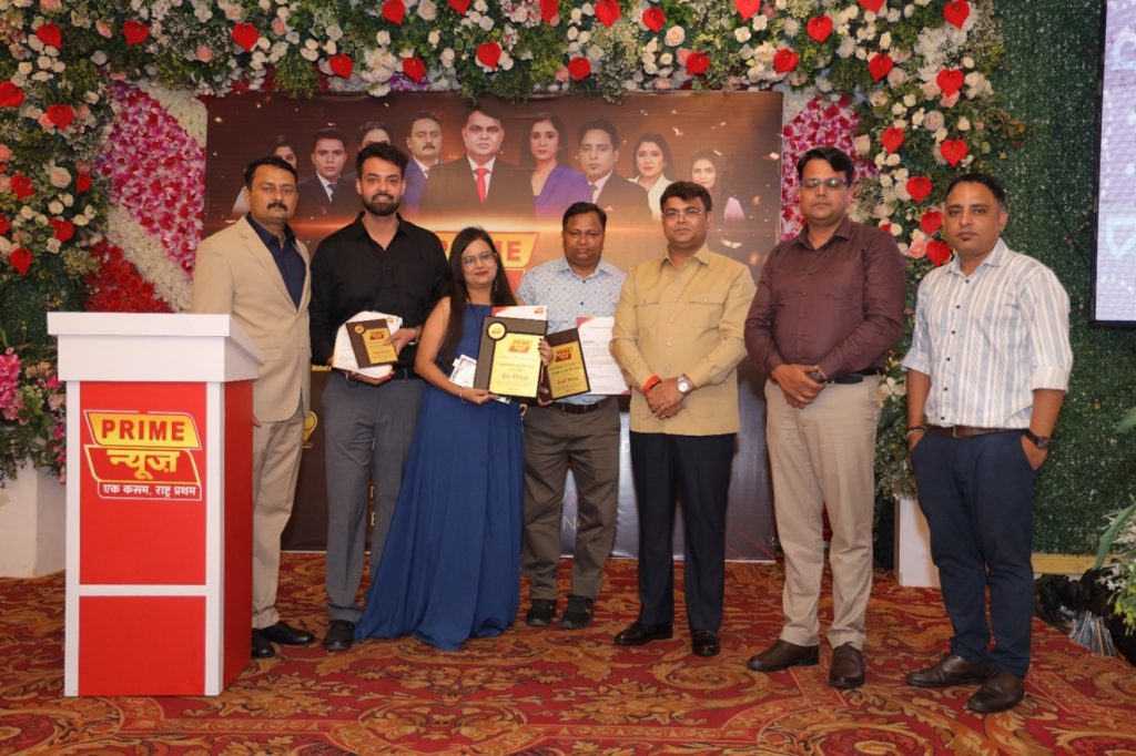 prime news_3 | No. 1 Indian Media News Portal Seven people on a decorated stage during an award ceremony; two recipients (a woman in a blue gown and a man) hold plaques while others stand beside them in formal attire with a 'Prime News' backdrop and floral arch.