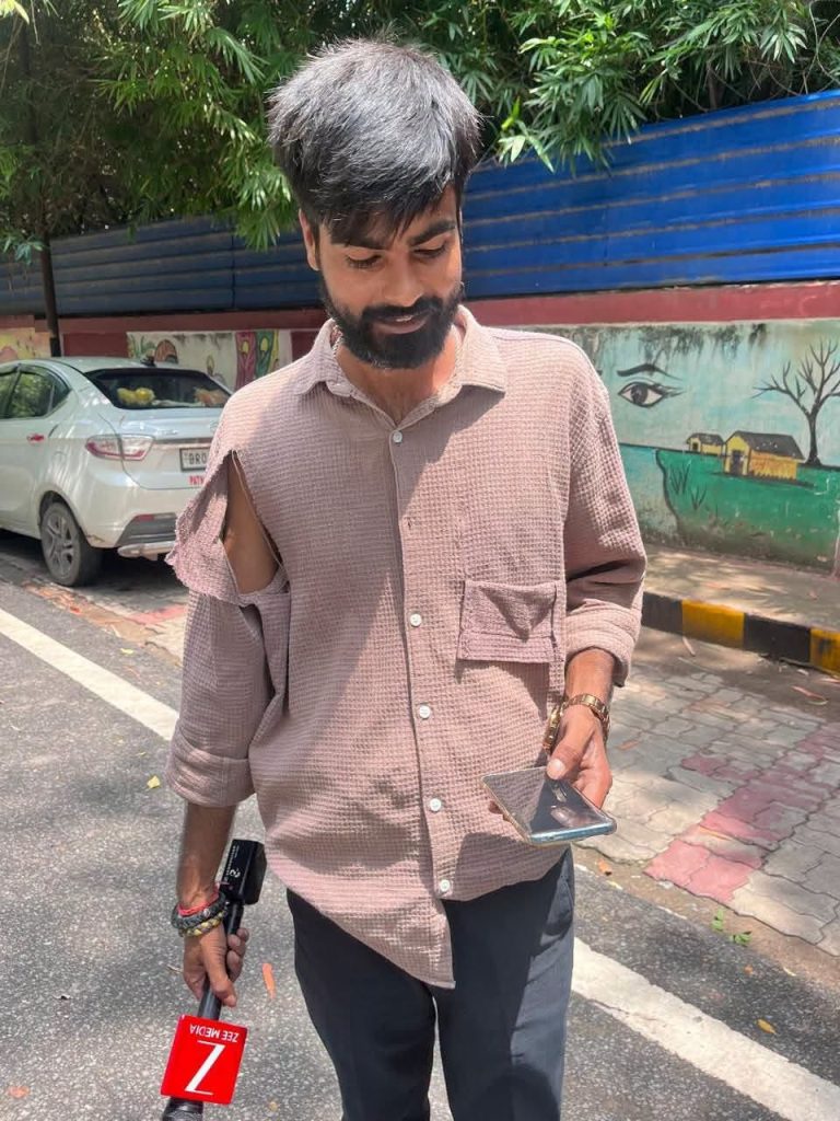 prince_zee | No. 1 Indian Media News Portal Man with a beard and torn-sleeve shirt looking down at his phone on a street with a reporter’s red Z microphone nearby.