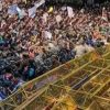 Dense crowd of protesters surrounding a yellow metal scaffold-like platform on a vehicle, in a chaotic public demonstration scene.